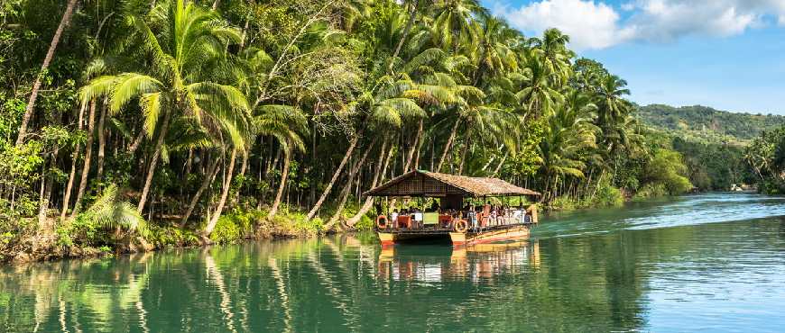 Loboc River in Bohol with calm green water, palm trees along the banks, and boats cruising through a tropical riverside village
