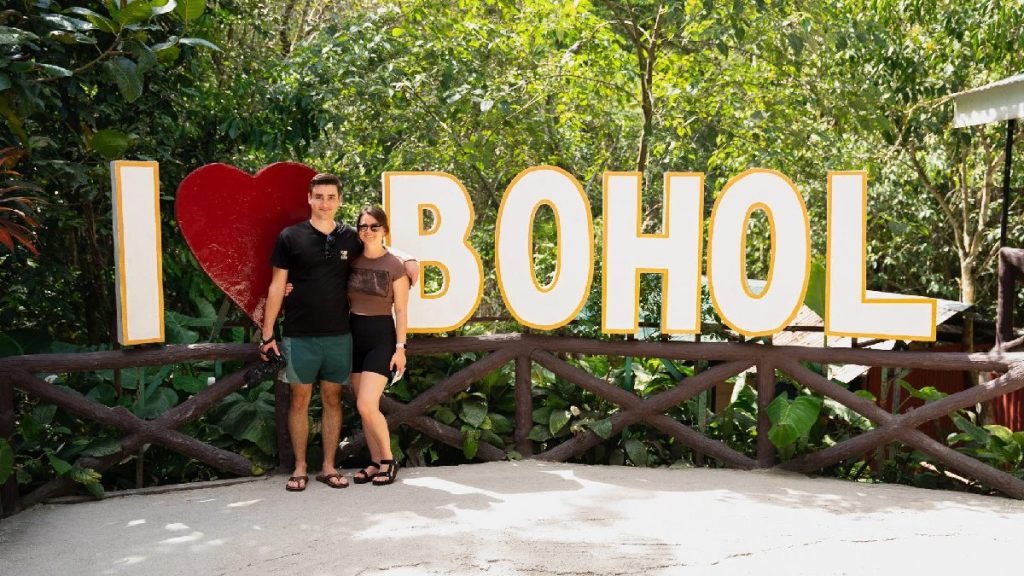 Tourists posing at the I Love Bohol sign surrounded by lush greenery, highlighting a Bohol tour package experience