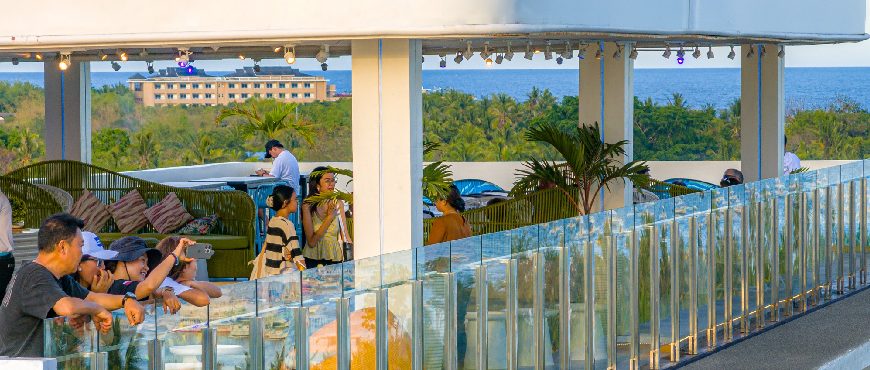 Rooftop lounge at Solea Bohol with guests relaxing by a glass railing, overlooking palm trees, resort buildings, and the ocean under a clear sky.