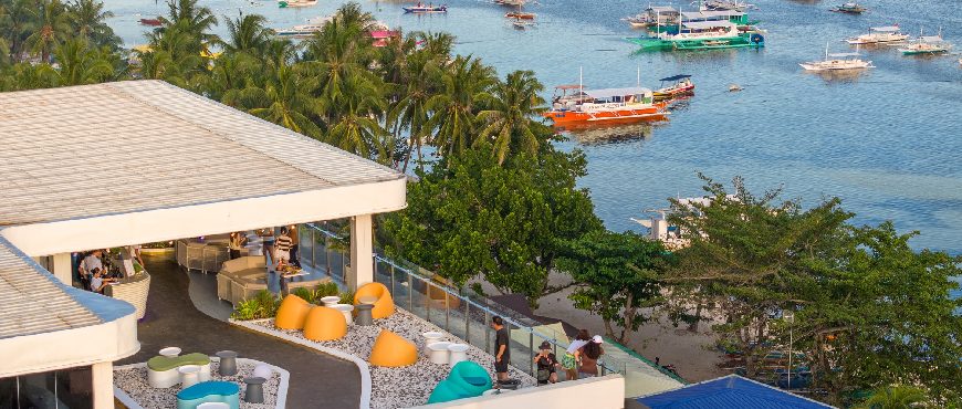Solea Bohol beachfront resort with modern terrace seating, palm trees, and boats anchored in the clear blue sea.